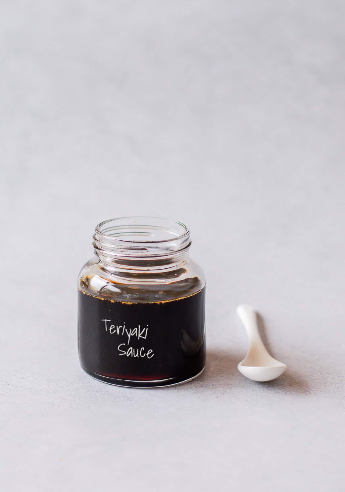 A small glass jar filled with dark teriyaki sauce, labelled Teriyaki Sauce, with a small white spoon resting beside it.