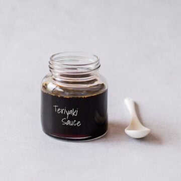 A small glass jar filled with dark teriyaki sauce, labelled Teriyaki Sauce, with a small white spoon resting beside it.