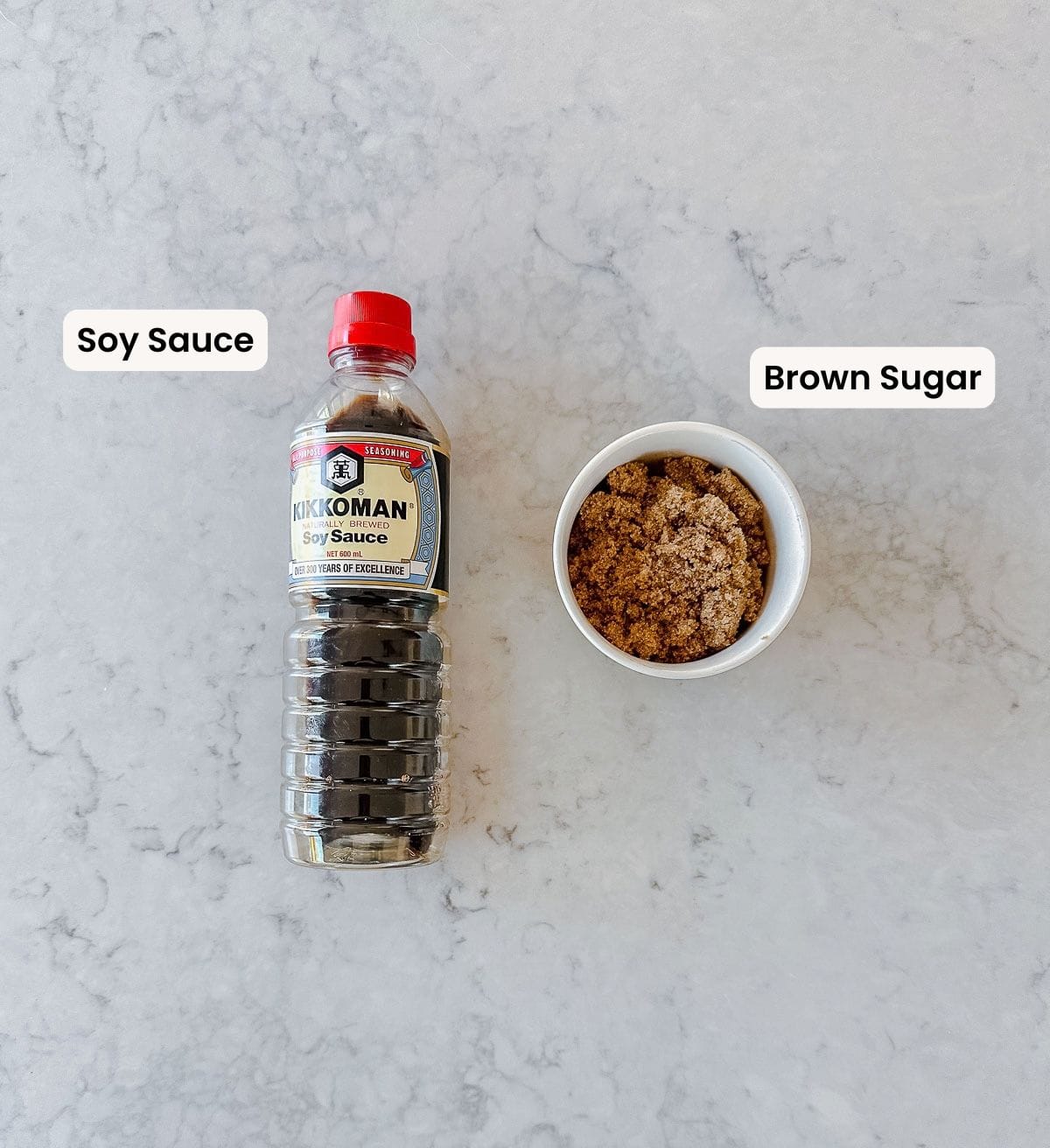 Overhead image of a bottle of soy sauce and a white bowl filled with brown sugar. Both labelled and on a white marble surface.