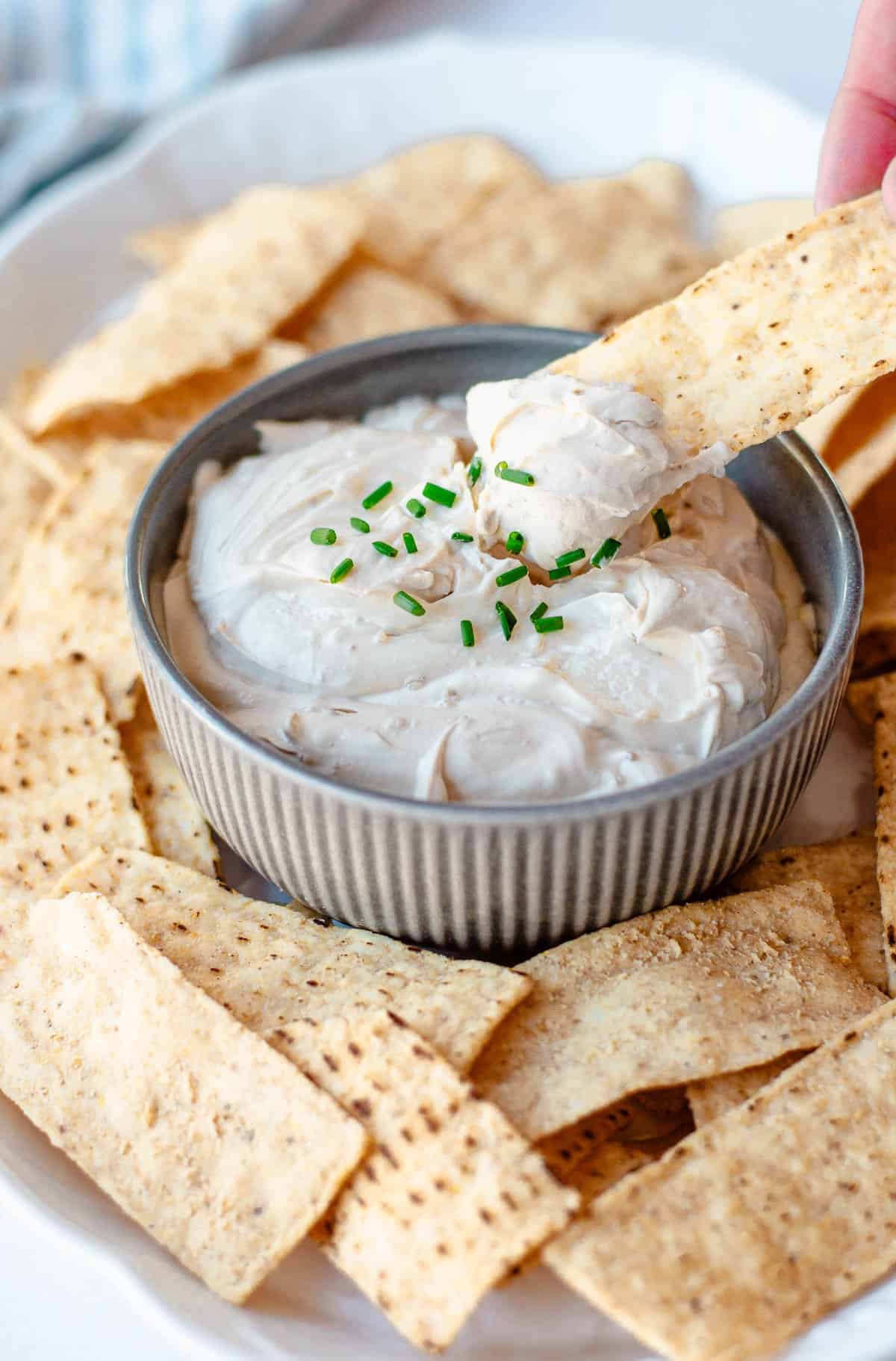 A hand dips a tortilla chip into a bowl of creamy dip topped with chopped chives, surrounded by more crisps.