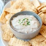 A small bowl of creamy dip topped with chopped chives, surrounded by toasted pitta chips on a platter.