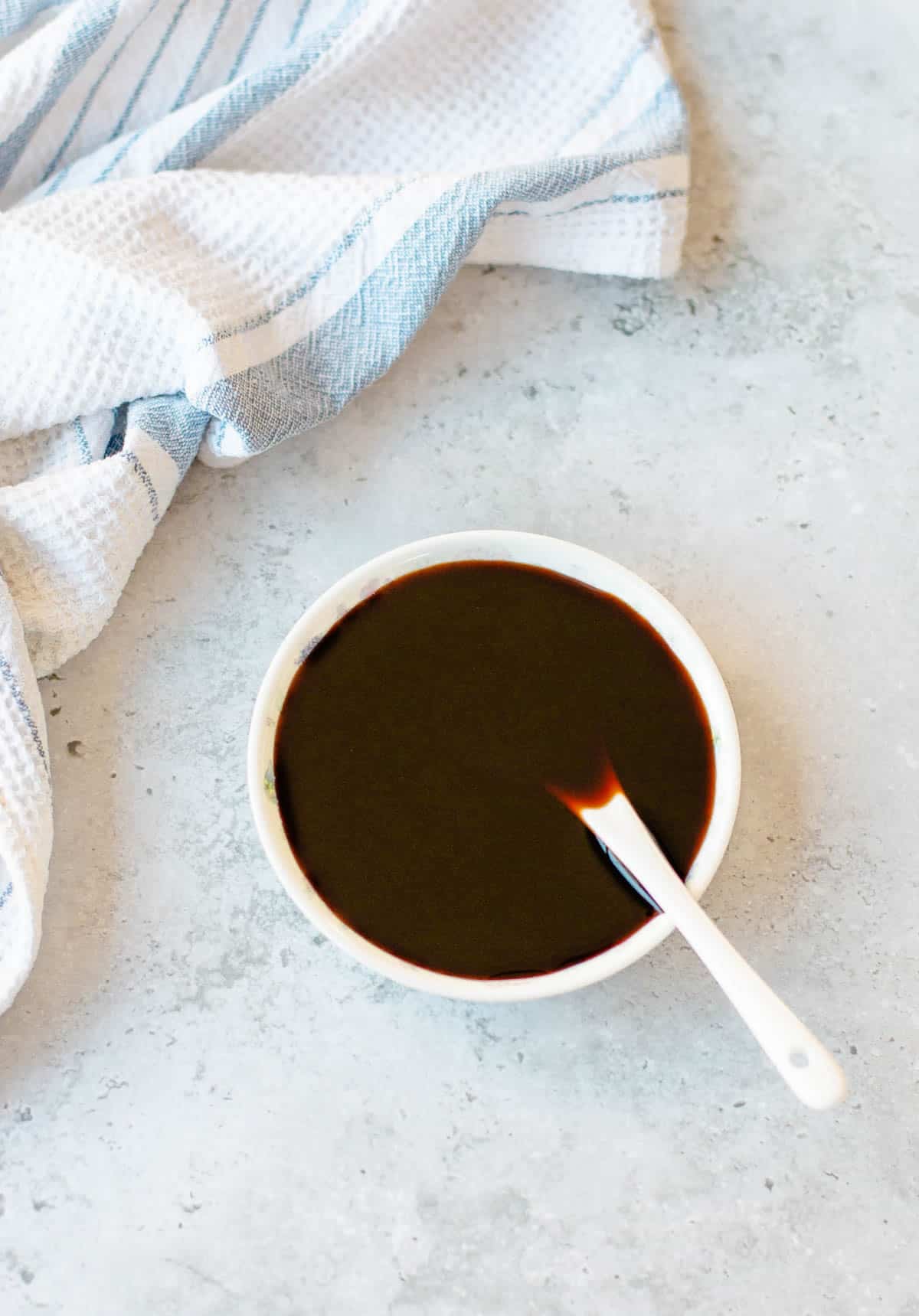 A small white bowl of kecap manis with a white spoon inside, set next to a white and blue striped kitchen tea towel.