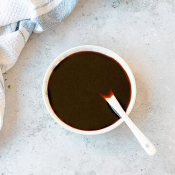 A small white bowl of kecap manis with a white spoon inside, set next to a white and blue striped kitchen tea towel.
