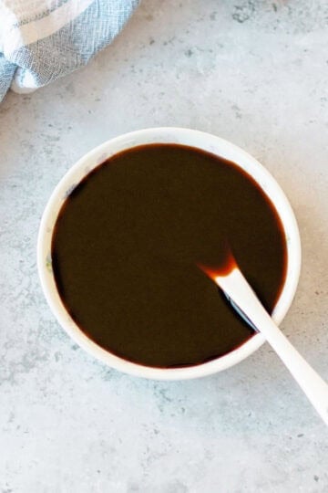 A small white bowl of kecap manis with a white spoon inside, set next to a white and blue striped kitchen tea towel.