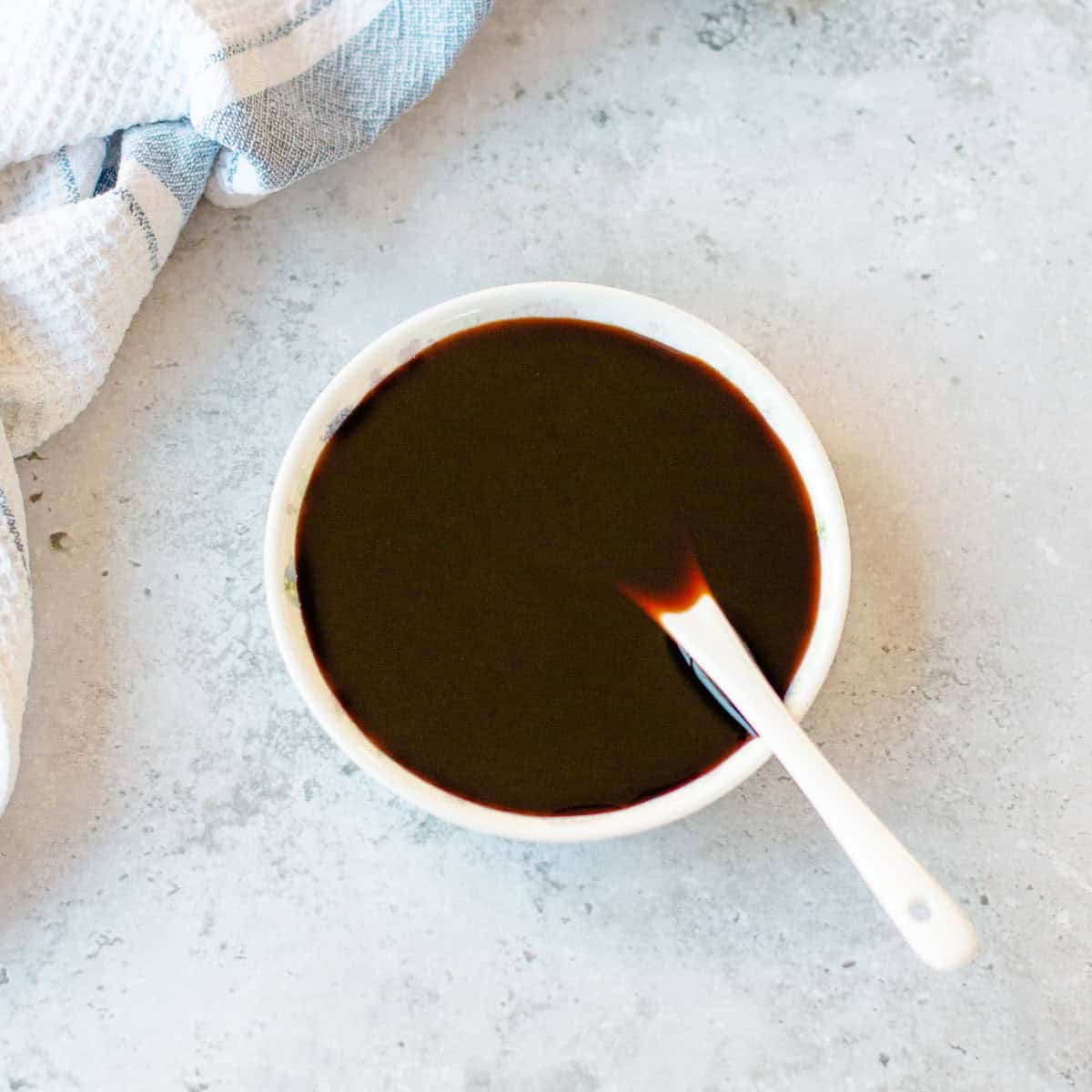 A small white bowl of kecap manis with a white spoon inside, set next to a white and blue striped kitchen tea towel.